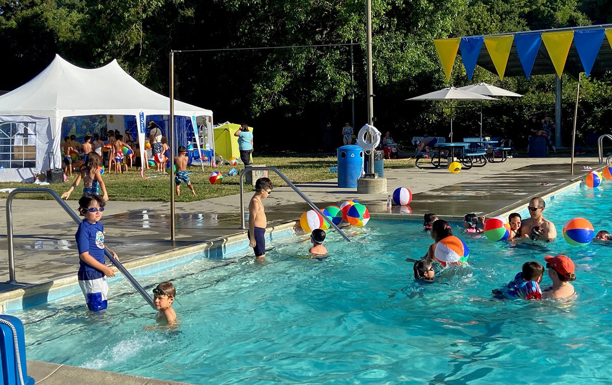 Kids swimming in the pool on a sunny day with covered picnic tables and tents