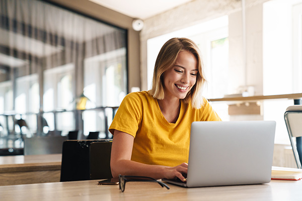 Photo of joyful nice woman using laptop and smiling while sitting at table in open-plan office