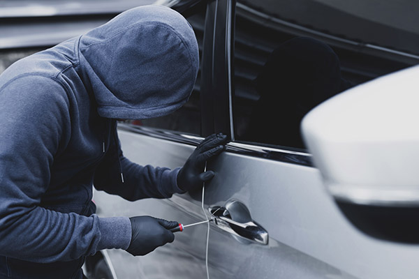 Thief wearing gloves and a hoodie uses screwdriver on door handle of car.