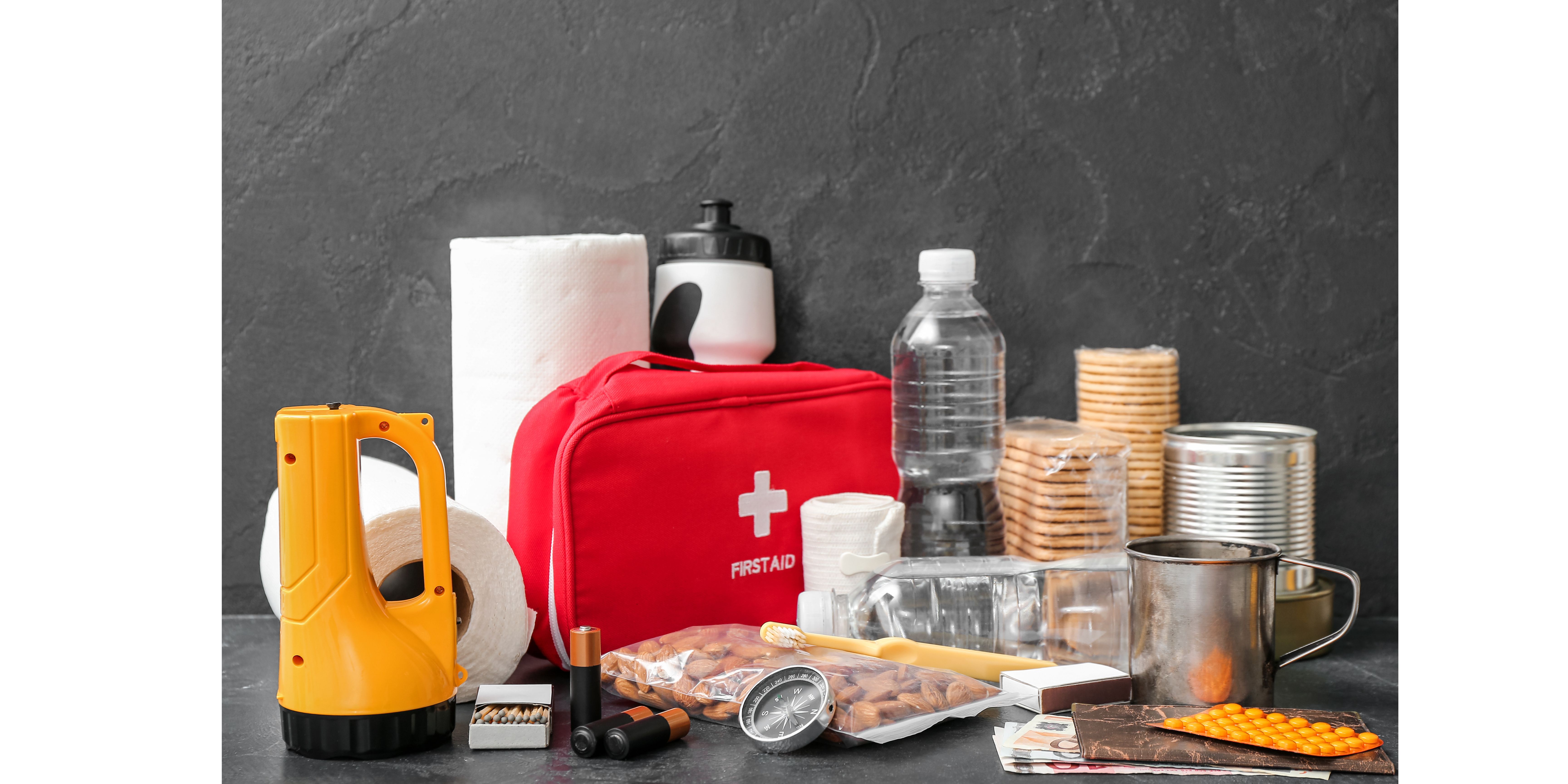 Emergency preparedness items (flashlight, first aid kit, water and other supplies) on a table with a dark grey wall.