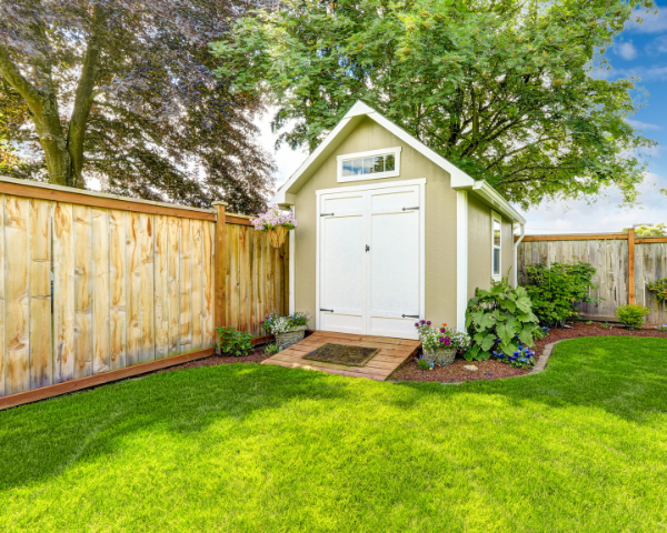 Detached residential unit in a backyard with dark siding, a window, adjacent landscaping, and a fenced property boundary.