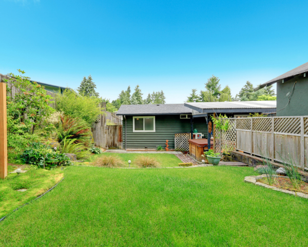 Small detached structure in a landscaped backyard, with white double doors, a gable roof, surrounding plants, and a wooden fence.