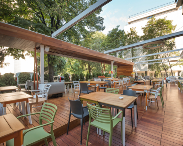 Outdoor dining area with wooden deck flooring, multiple tables and chairs, overhead shade structures, and surrounding trees and landscaping.