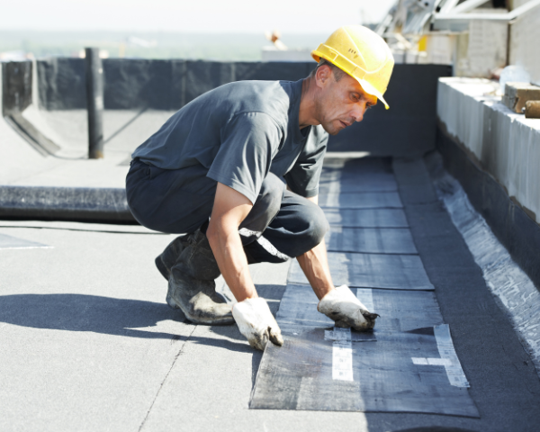 An individual installing roofing material on a flat roof, placing rectangular sections of membrane along the surface with tools and equipment nearby.