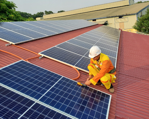 Worker performing installation work on solar panels mounted across a large metal roof at a commercial or industrial building, using a tool while positioned beside multiple rows of photovoltaic panels.