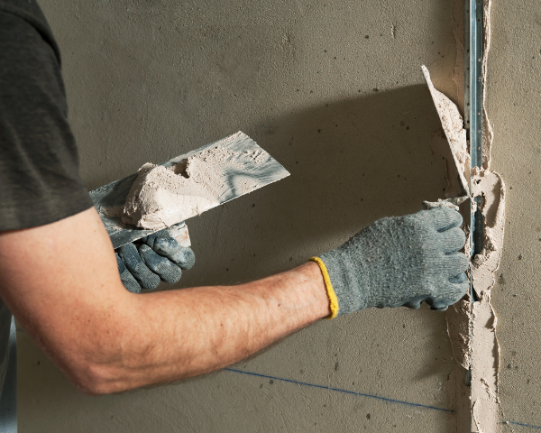 An individual applying plaster or joint compound to a vertical surface using a metal trowel, holding additional compound on a second tool.