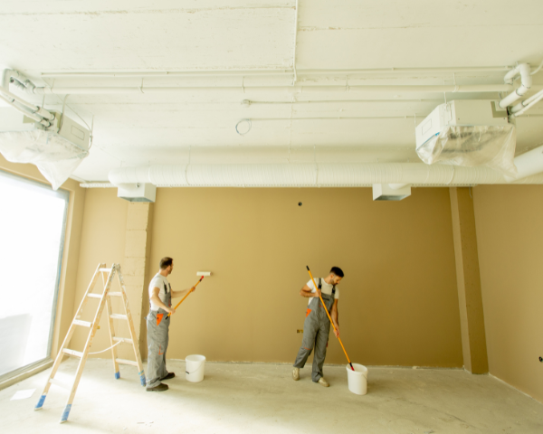 Two individuals painting a large interior wall using rollers, with buckets of paint, a ladder, and exposed ceiling ductwork visible.