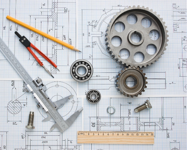 Mechanical components and drafting tools arranged on top of technical engineering drawings, including gears, bearings, a caliper, a compass, bolts, a pencil, and a ruler.