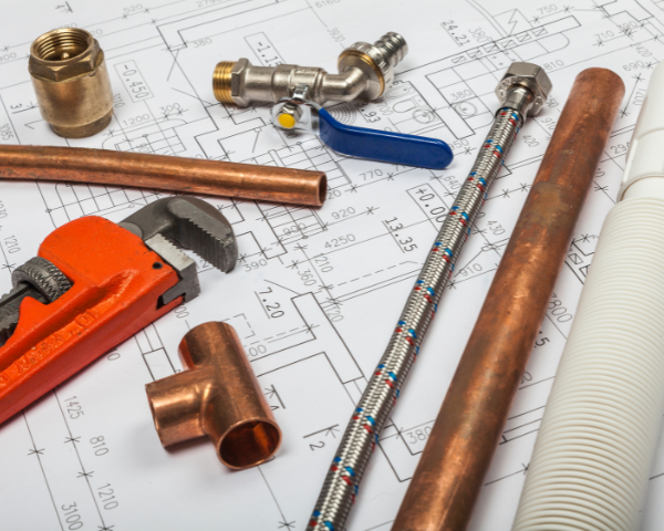 Plumbing tools and copper pipe fittings placed on top of technical plumbing drawings, including a pipe wrench, copper pipes, connectors, and a valve.