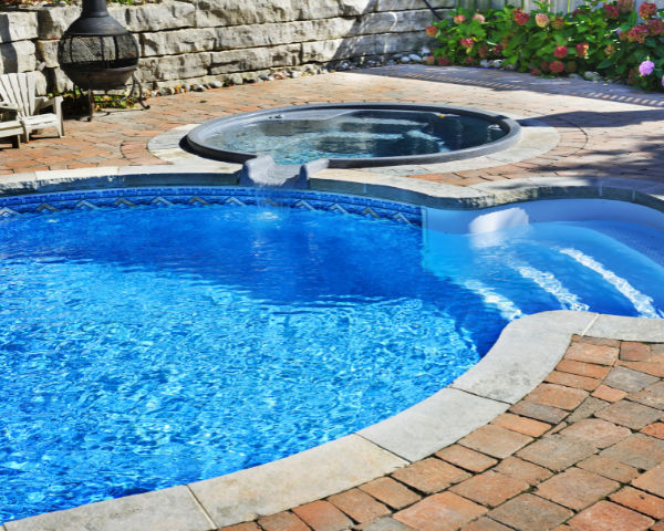 Outdoor swimming pool with clear blue water next to a raised circular spa, surrounded by brick pavers, landscaping, and a stone retaining wall.