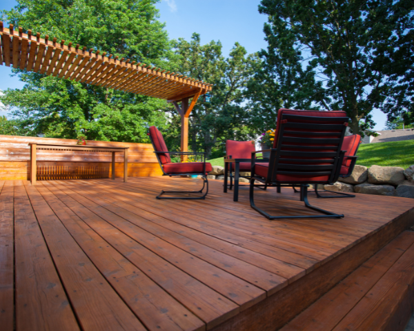 A wooden outdoor deck with a pergola overhead, furnished with red cushioned chairs and a table, surrounded by trees and landscaping.