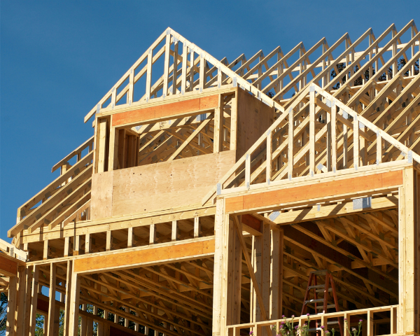 Wood framing of a new residential structure under construction, showing exposed studs, beams, trusses, and an unfinished upper story against a clear sky.