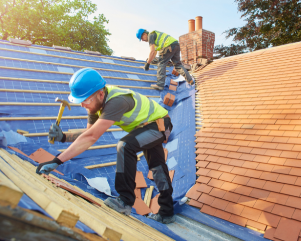 Workers installing new roofing materials on a pitched roof, placing wooden battens and tiles over a blue underlayment.