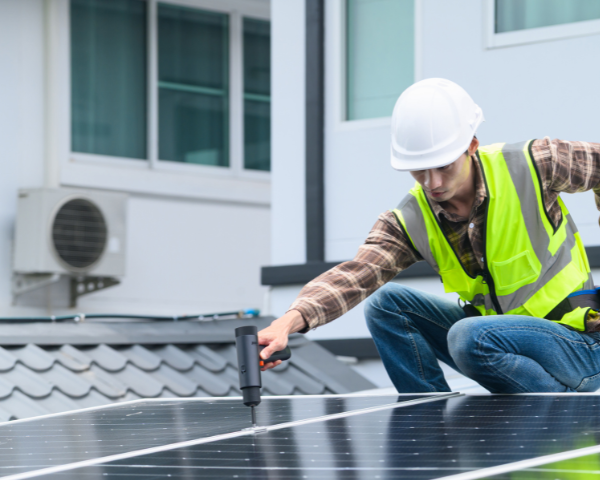 Worker installing or adjusting rooftop solar panels on a residential building, using a handheld tool while sitting on the roof near dark reflective photovoltaic panels.