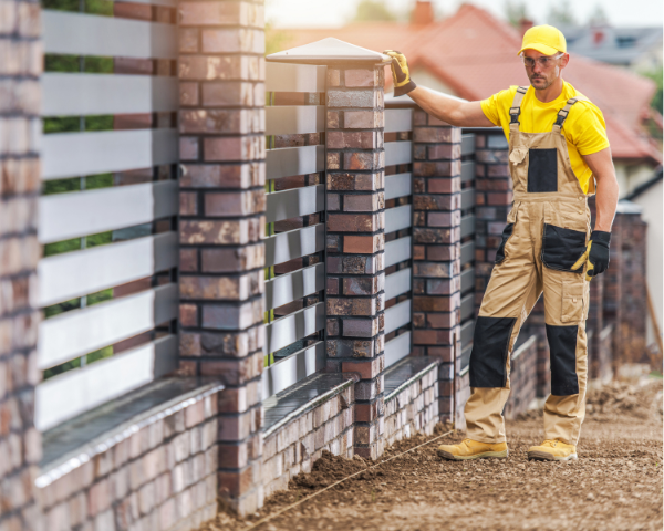 A worker standing next to a newly constructed fence made of alternating horizontal metal slats and brick columns.