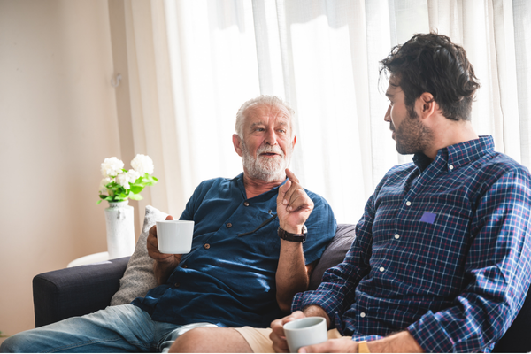 “Two people sitting on a sofa in a bright living room, each holding a mug, engaged in conversation. A small plant in a white pot sits on a table behind them, and sheer curtains allow soft natural light into the space.