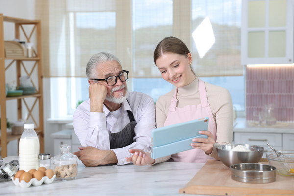 Two people standing at a kitchen counter looking at a tablet together, with baking ingredients and utensils in front of them