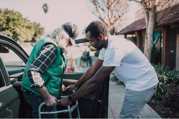 Two people are outdoors next to a parked car, with one person assisting another who is using a walker