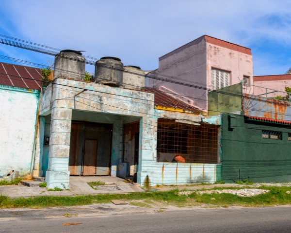 Weathered storefront and adjacent buildings with peeling paint, rusted metal bars, and water tanks on the roof along a roadside.