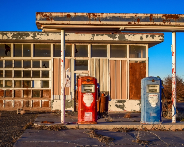 Abandoned gas station with two weathered fuel pumps in front of a deteriorating building with peeling paint and rusted metal.